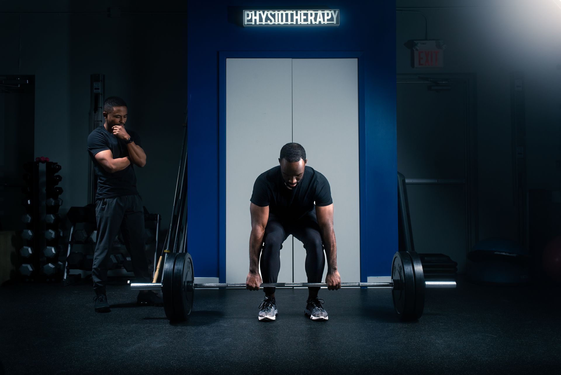 A person performs a barbell deadlift in a gym, while an observer watches them with their hand on their chin.