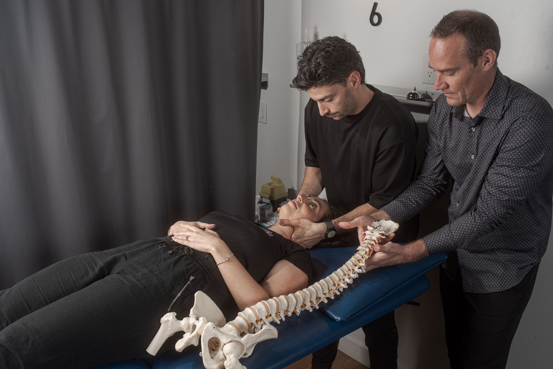 A patient lying on a treatment table receives a neck adjustment from a therapist, with a model spine shown for reference.