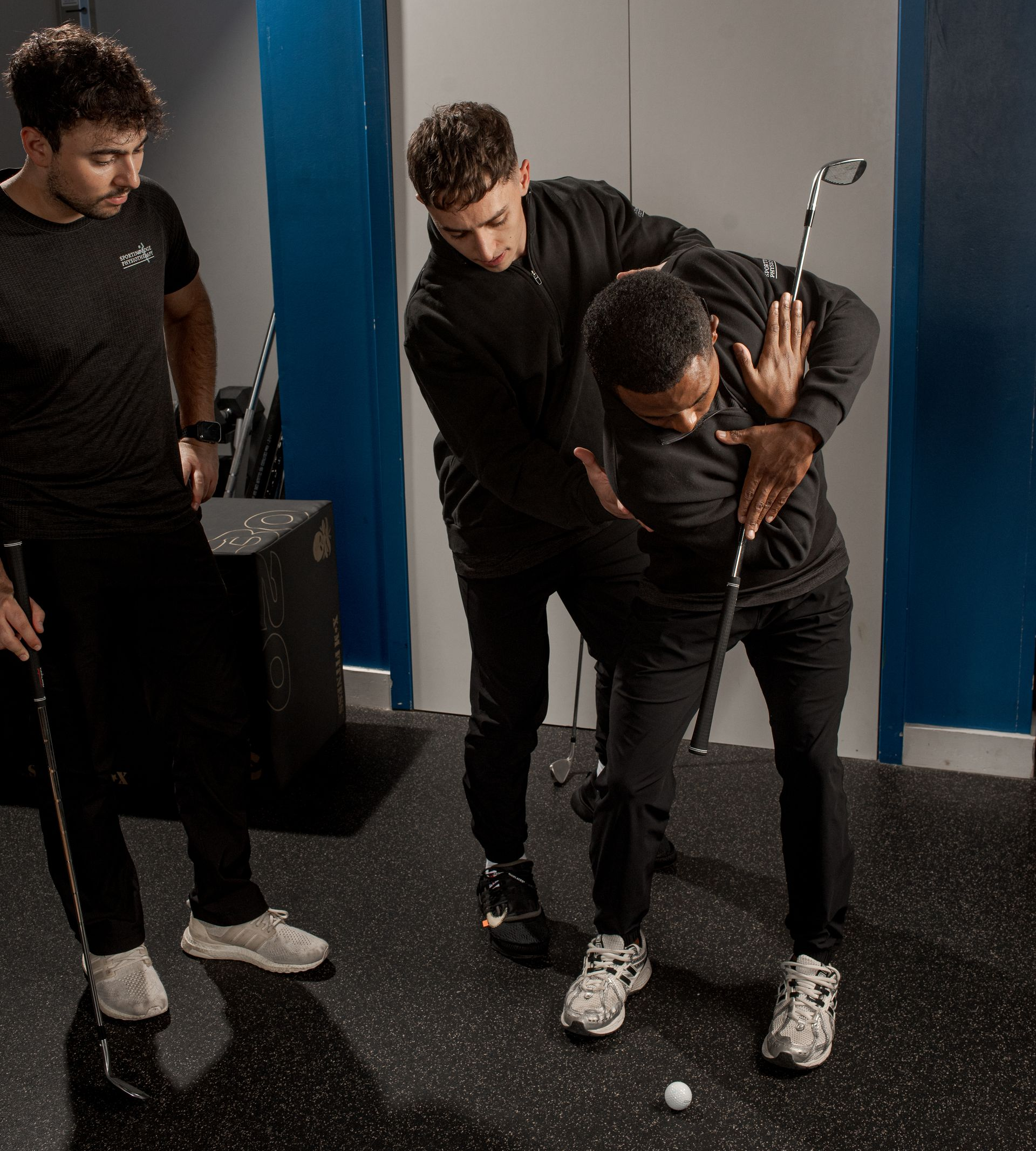 A coach guides a student in a golf stance, adjusting their posture while holding a club in an indoor gym setting.