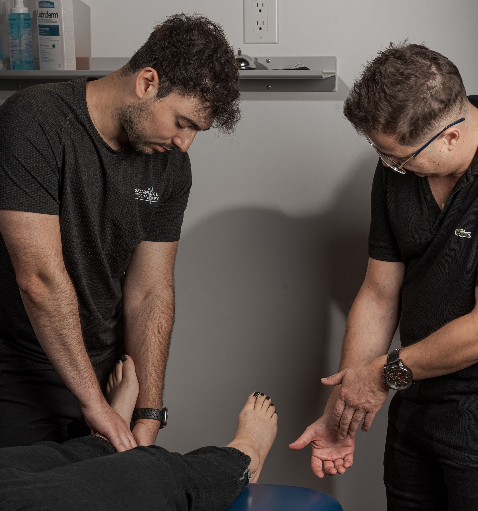Two practitioners in a clinic perform a physical assessment on a patient's leg resting on an examination table.