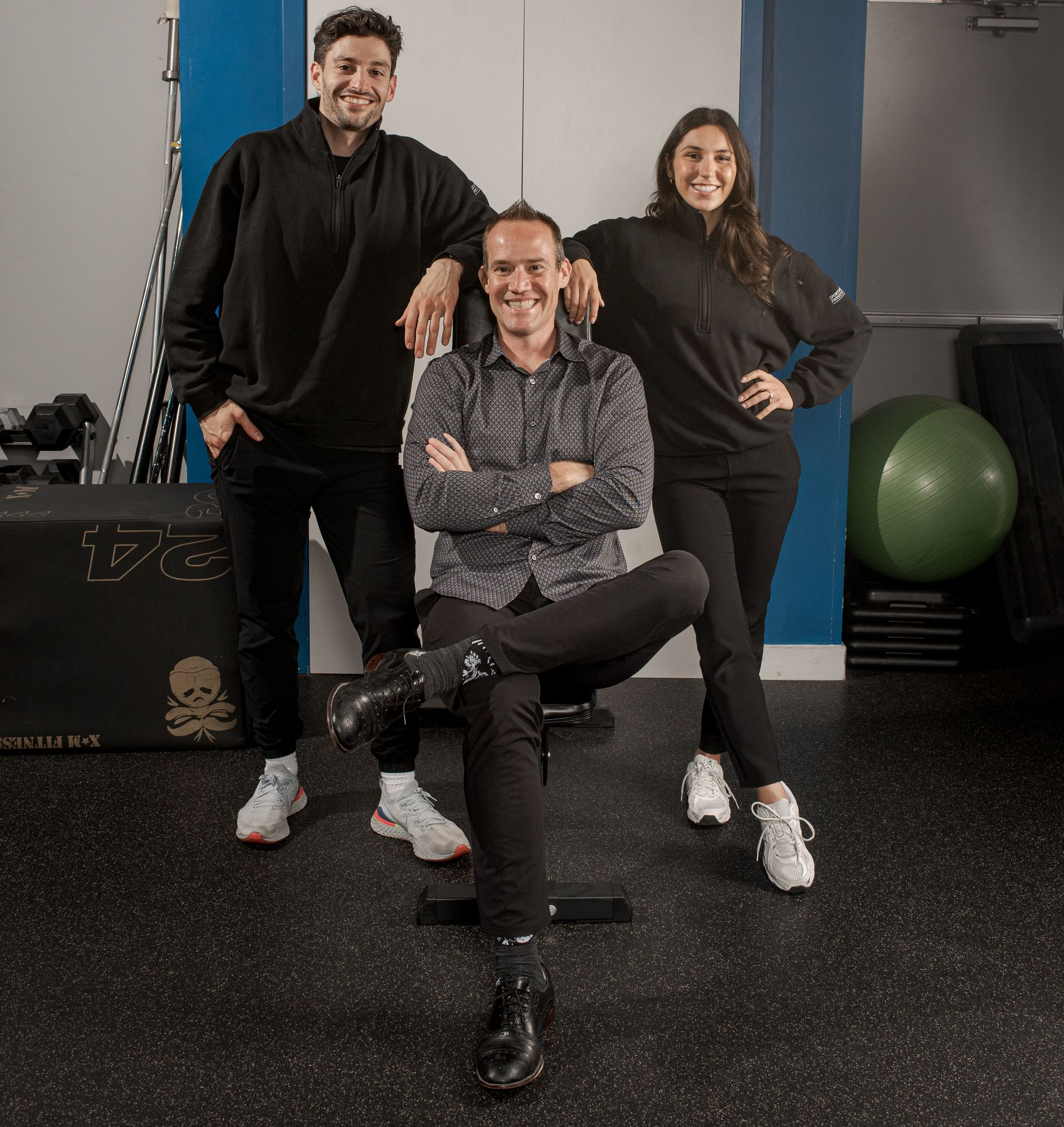 Three people pose in a gym; two stand behind a seated individual in a patterned shirt, all smiling toward the camera.