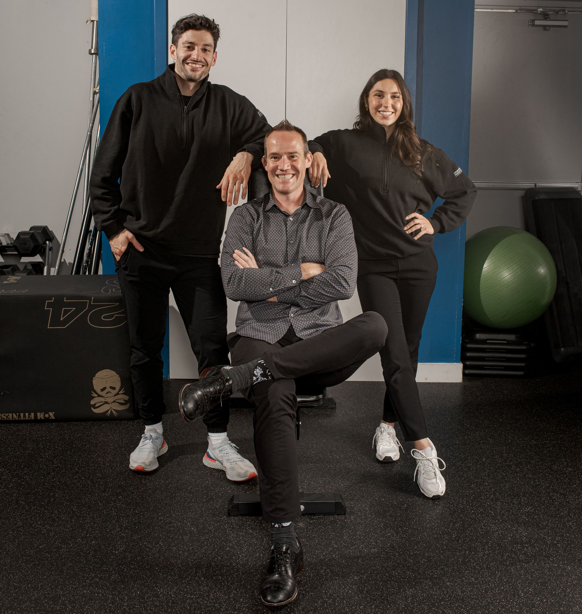 Three people pose for a photo in a physiotherapy clinic in front of a blue wall with a sign reading PHYSIOTHERAPY.