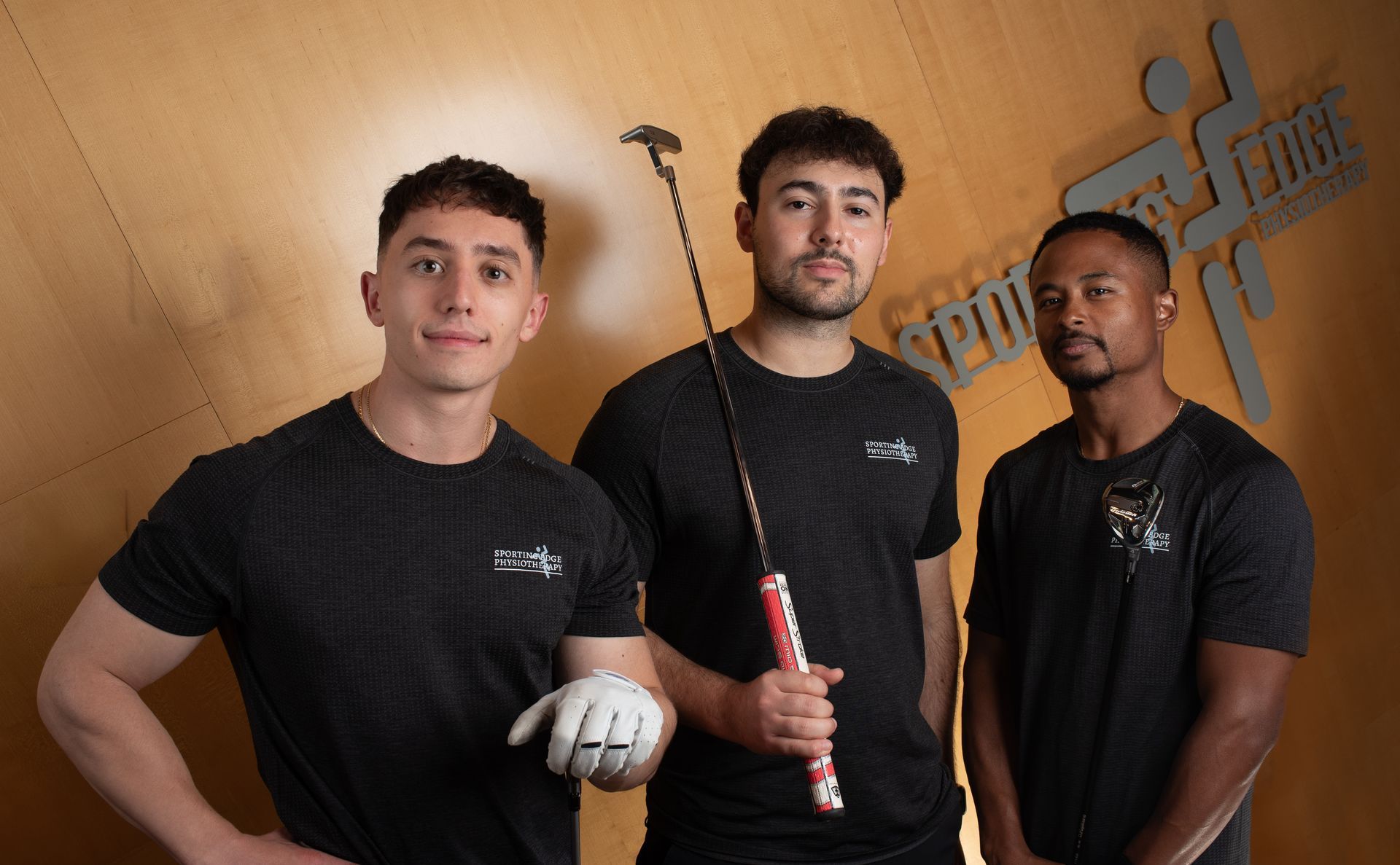 Three people in dark grey athletic shirts pose against a wooden wall; one holds a golf putter and another a white glove.