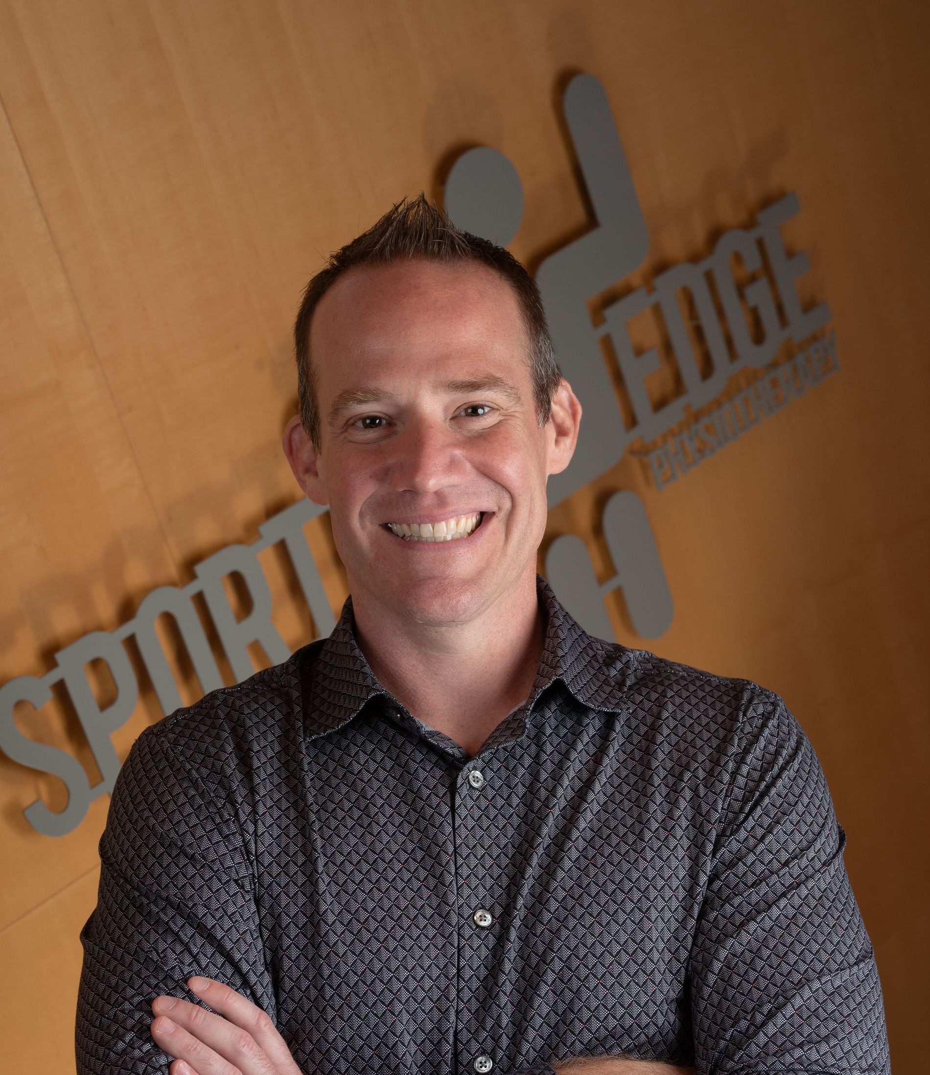 Matthew Wright A smiling person wearing a patterned button-down shirt, standing in front of a wall with the logo