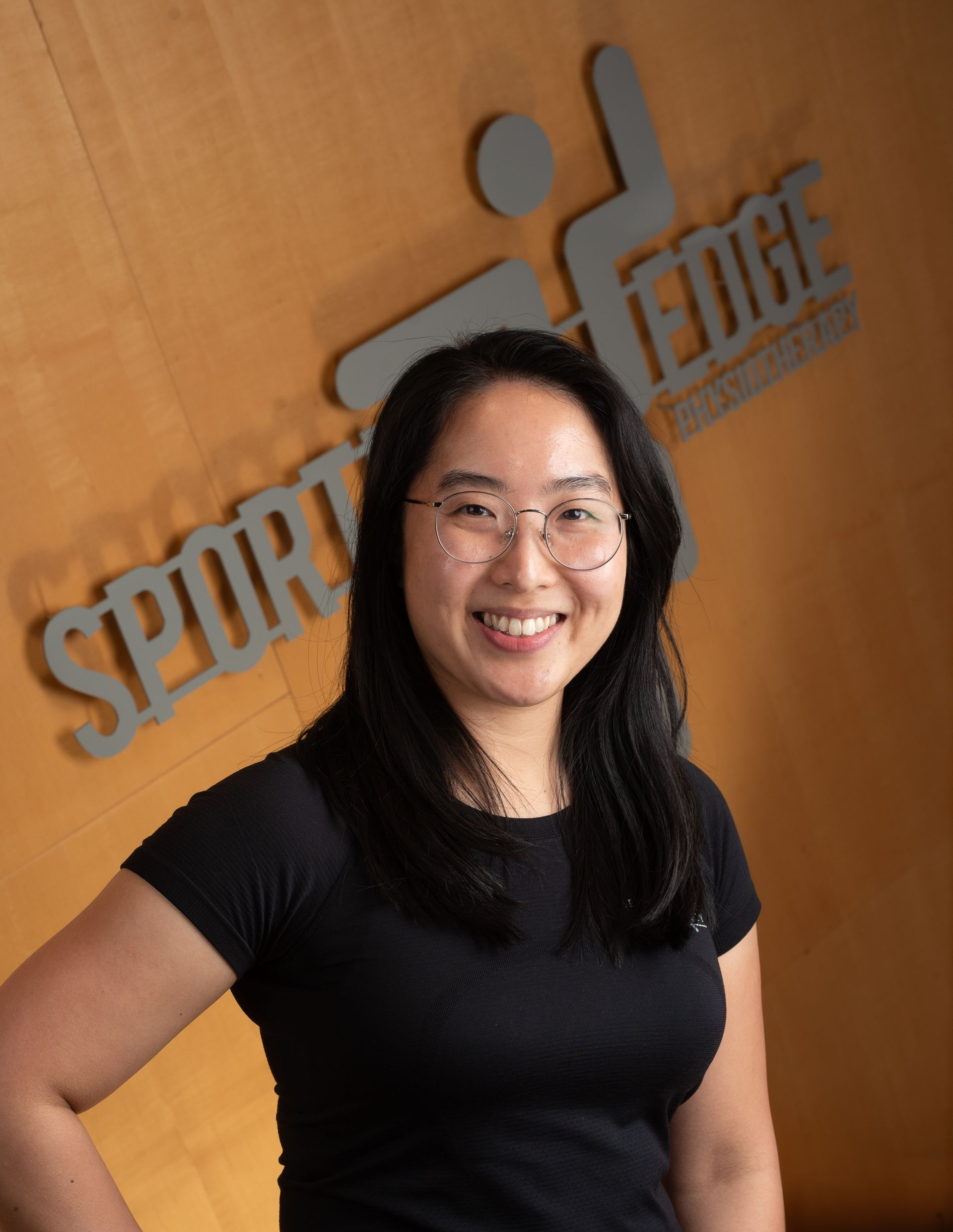 Kathleen Li A smiling person wearing glasses and a black shirt stands in front of a wooden wall with the Sporting Edge logo.