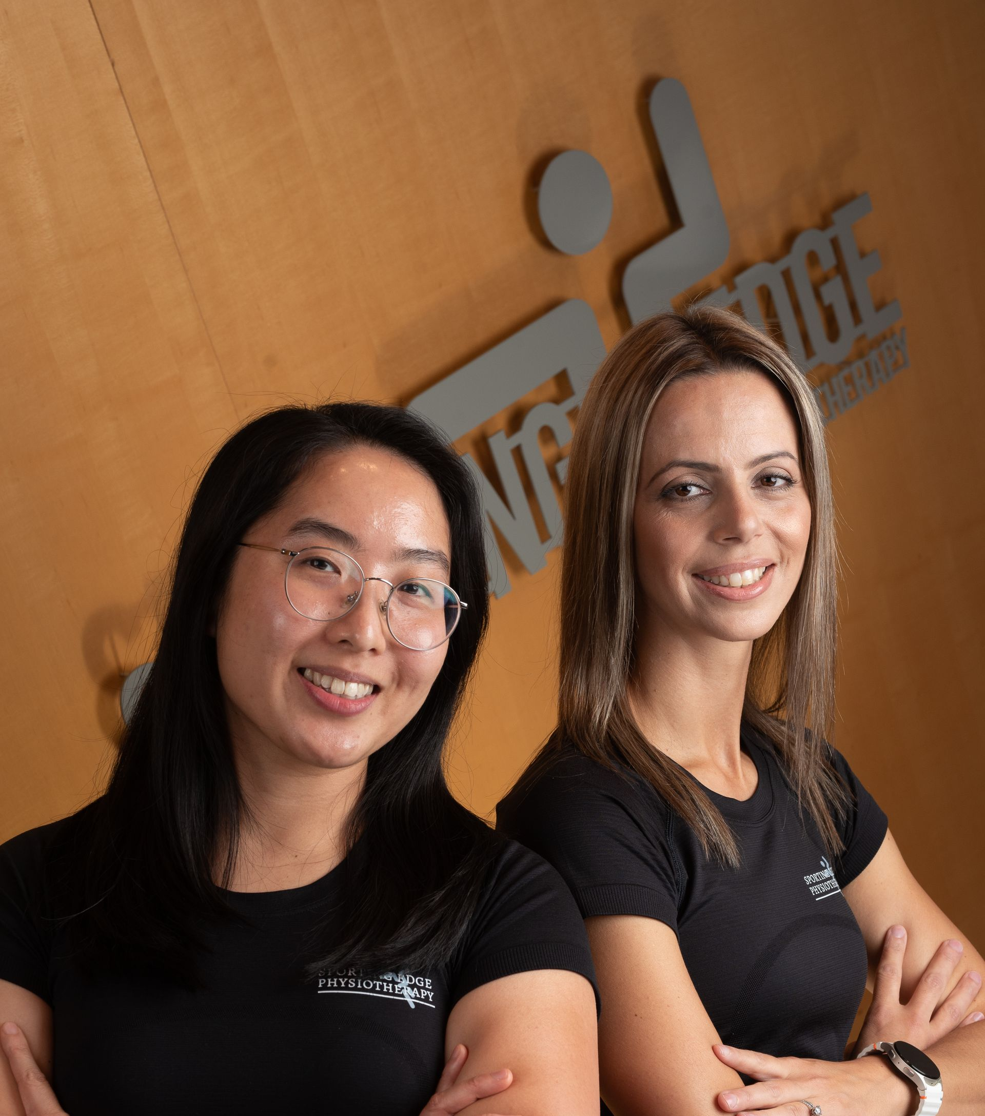 Two people stand side-by-side with arms crossed against a wooden wall with a partially visible corporate logo.