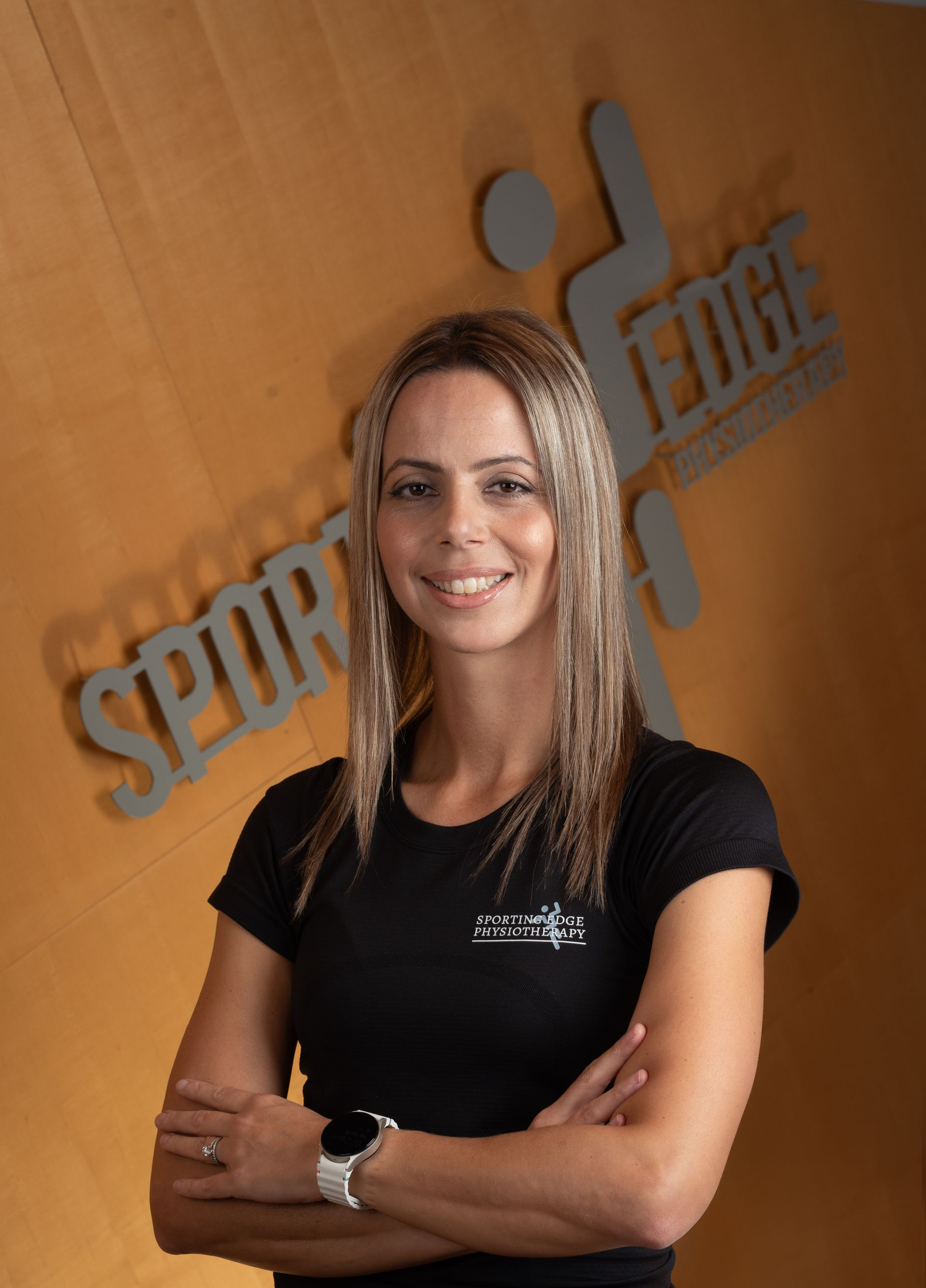 Marlene Luis A person with long highlighted hair stands with arms crossed against a wooden wall featuring a metallic Sporting Edge logo.