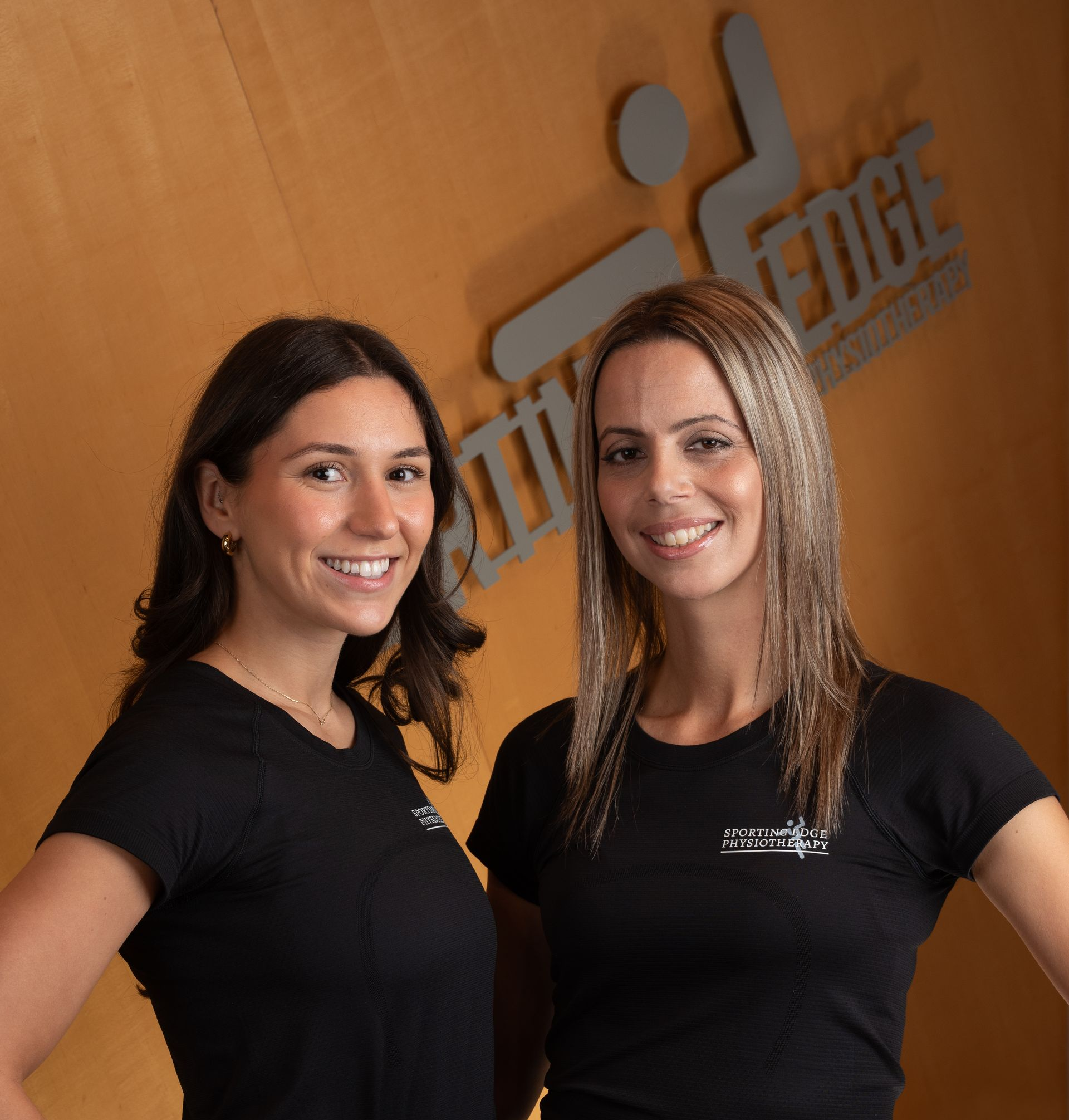 Two smiling professionals in matching black shirts pose in front of a wooden wall with a logo for Sporting Edge Physiotherapy.