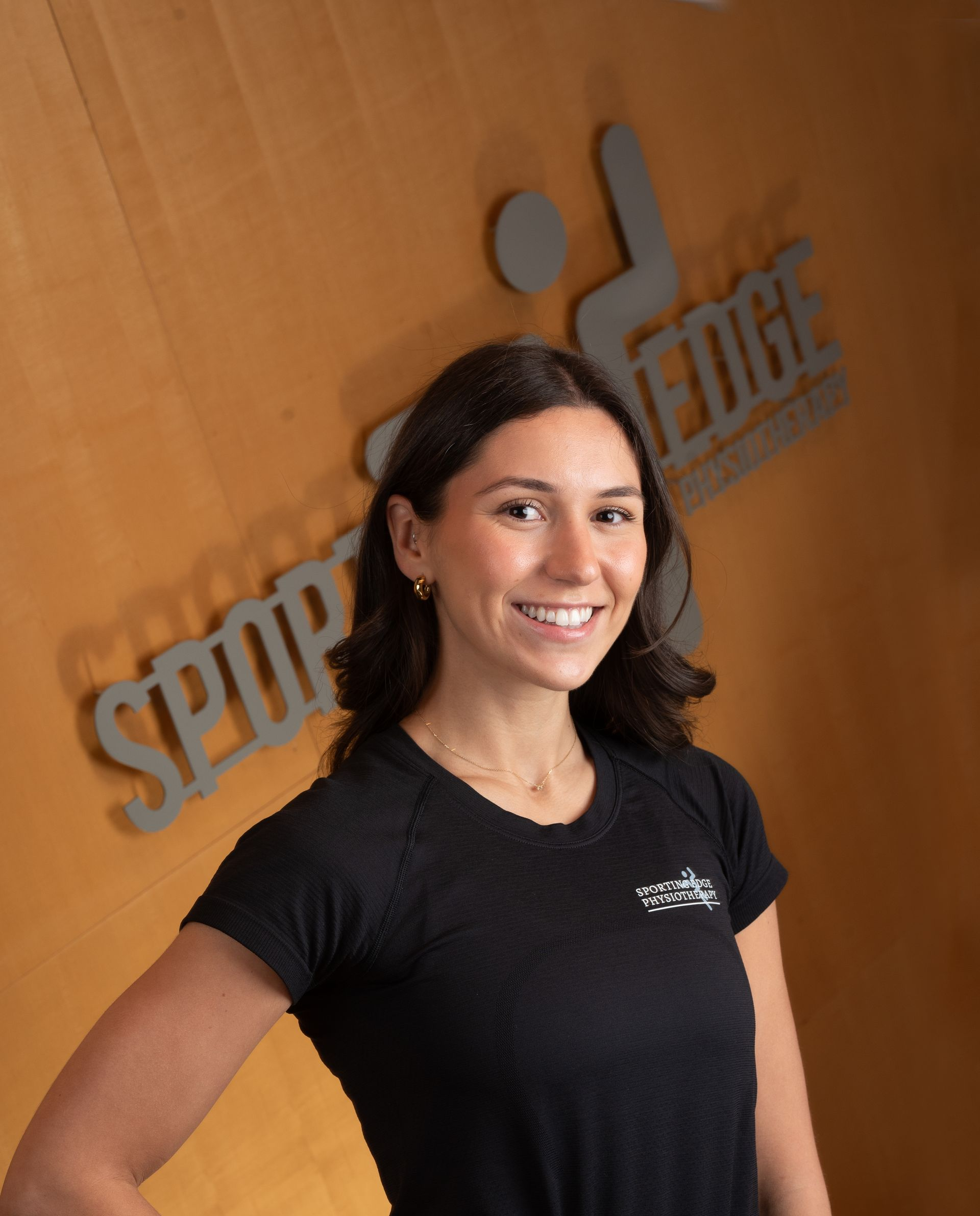 Dominique Tersigni A smiling woman in a black shirt standing in front of a wood-paneled wall with a grey Sporting Edge Physio logo