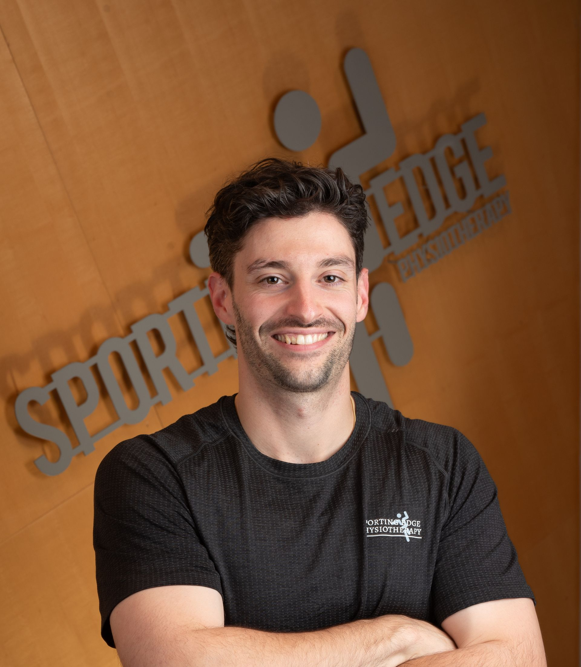 Marco Bandiera A person smiling with arms crossed in front of a wooden wall featuring a grey Sporting Edge Physio logo
