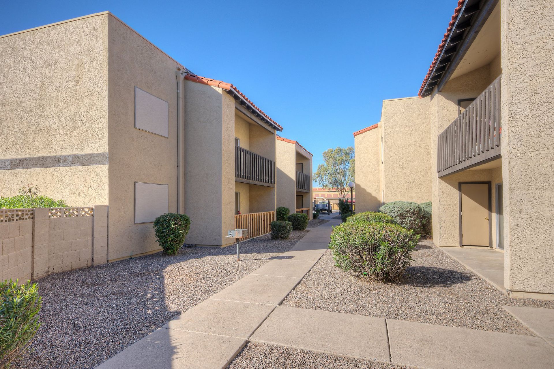 Exterior walkway between tan, stucco apartment buildings with desert landscaping under a clear blue sky.