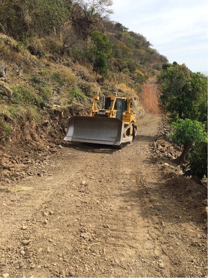 Bulldozer en un camino de tierra, despejando una ladera. Máquina amarilla, camino marrón, árboles verdes.