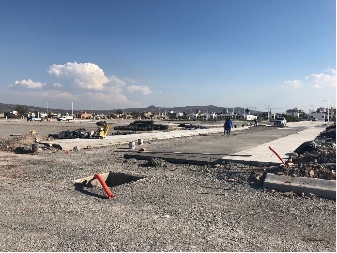 Zona en construcción con grava, hormigón y tuberías rojas. Una persona camina por la carretera.