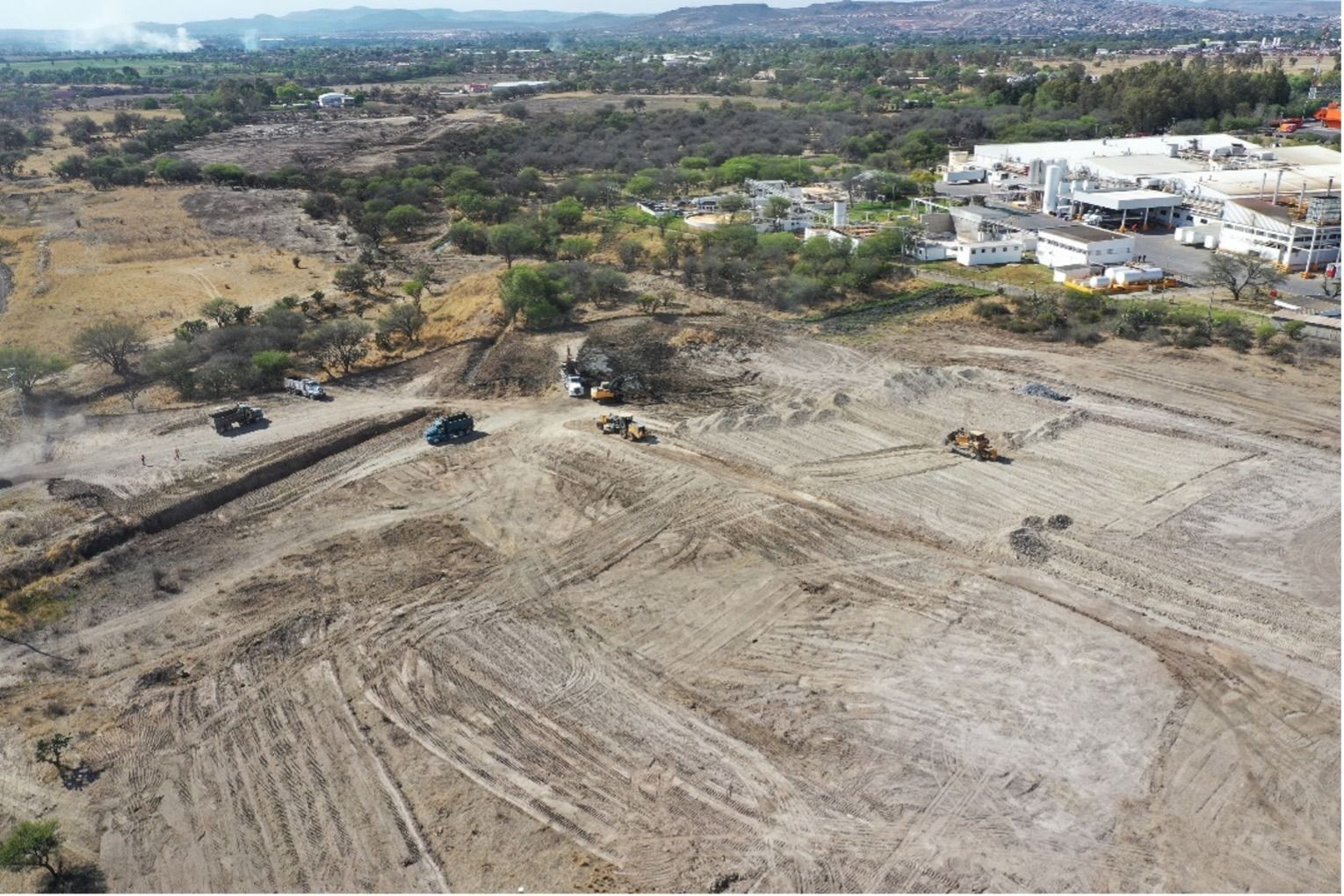 Zona de construcción con excavadoras limpiando el terreno cerca de un complejo de edificios y colinas a lo lejos.
