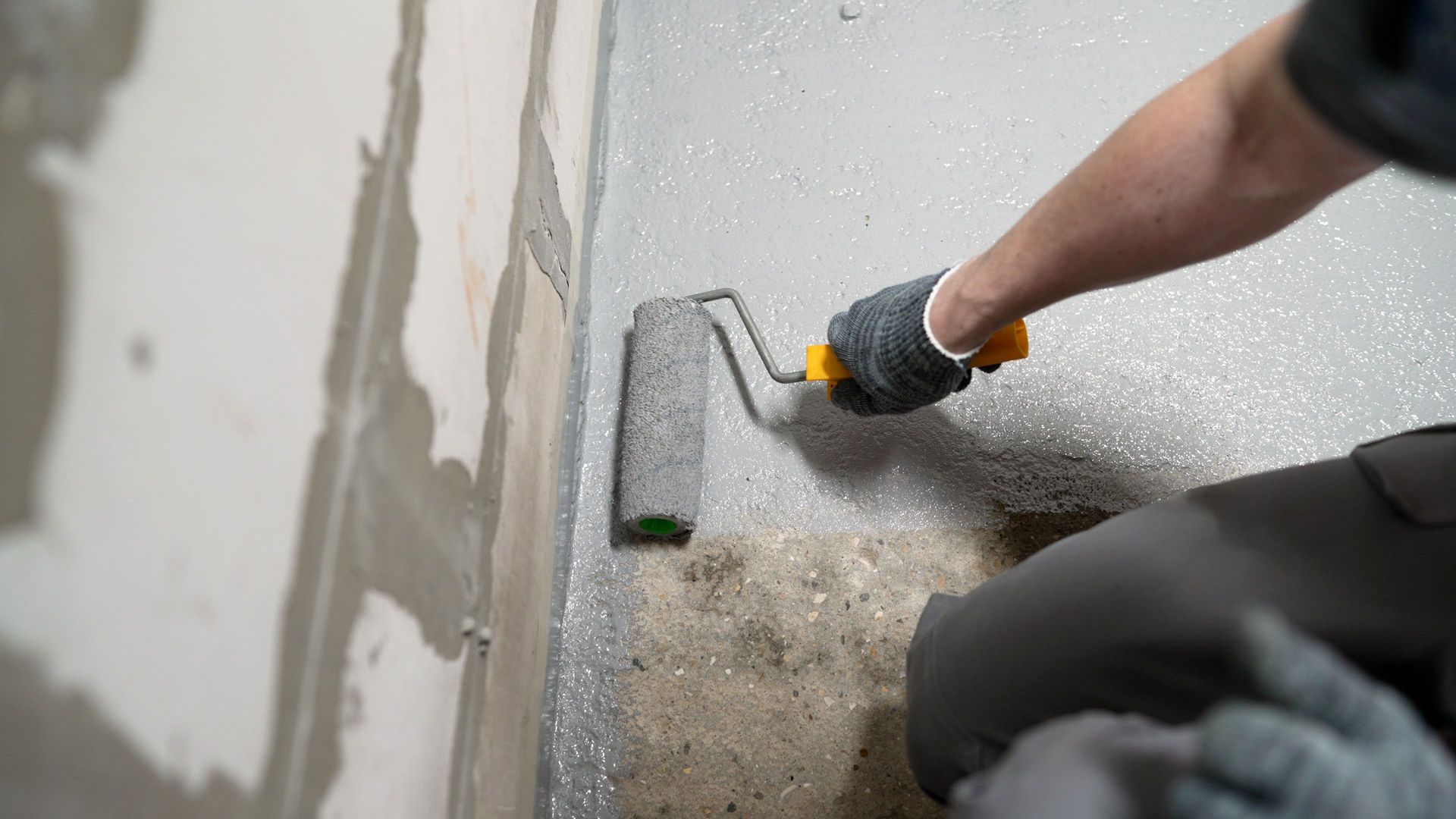 Gloved hand using a tool to scrape or repair a gray wall corner on a concrete floor