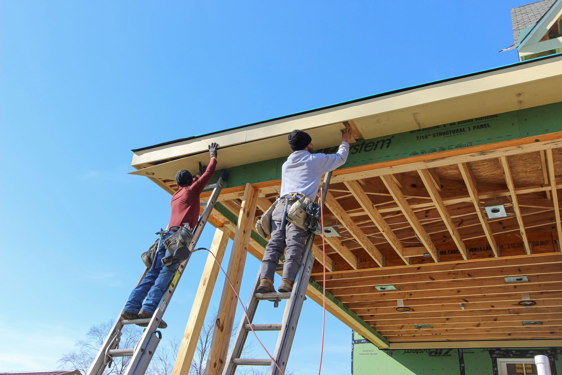 Two workers on ladders installing trim under a house roof on a sunny day at redwood roofing and exteriors