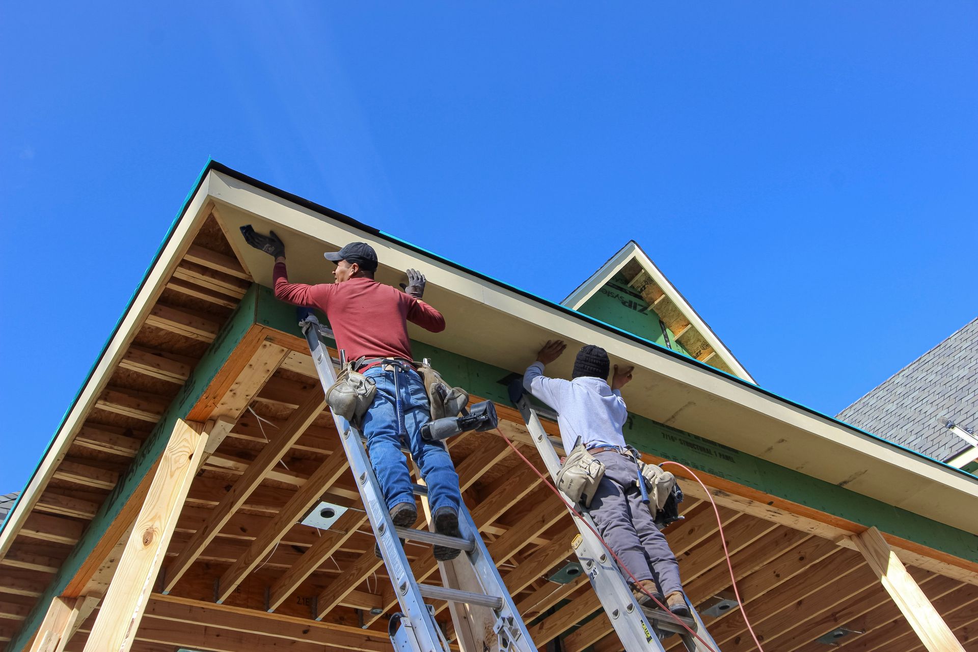 Two workers installing siding on a house under construction, viewed from below against a blue sky at redwood roofing and exteriors