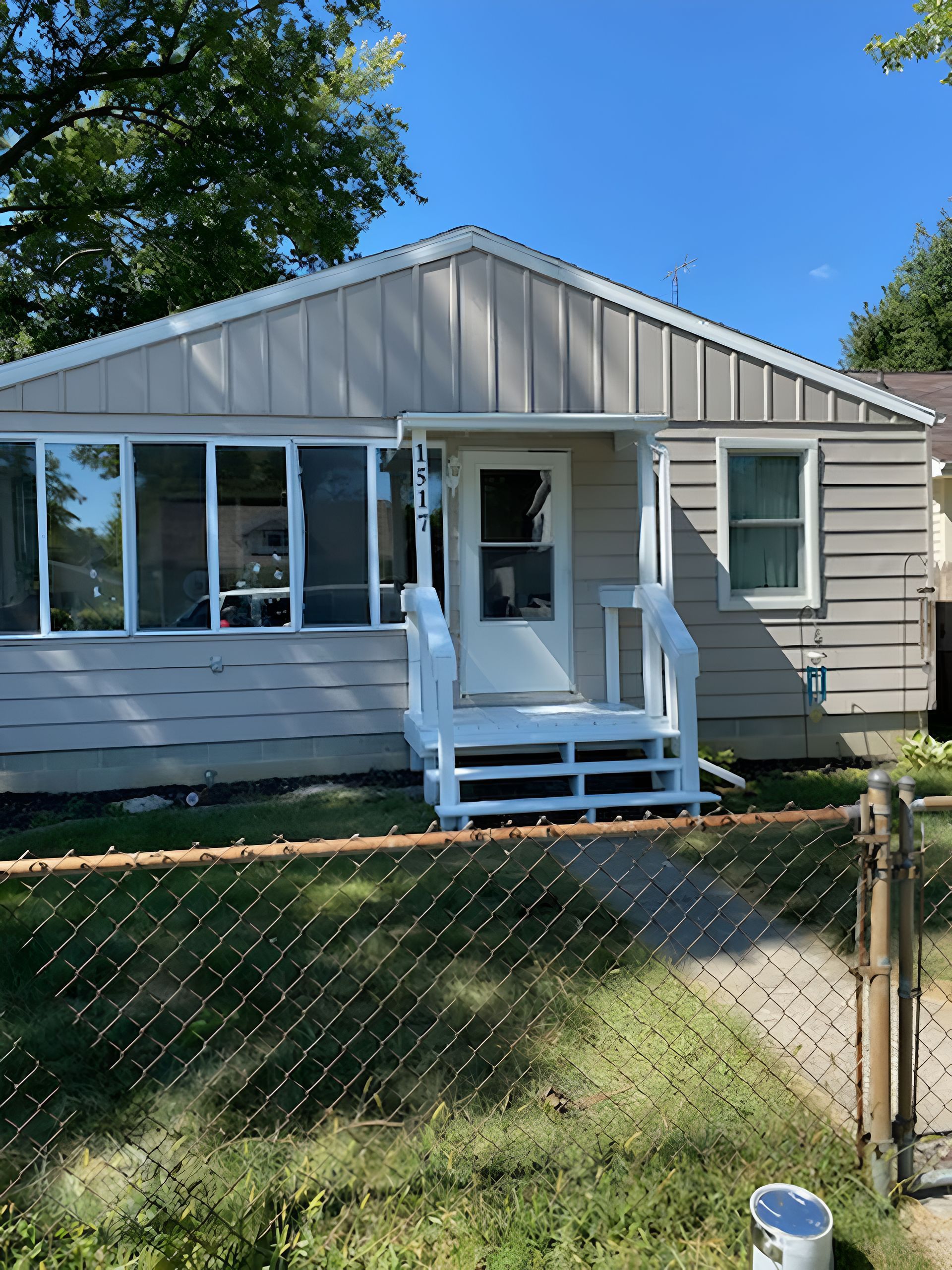 A beige single-story house with a white front porch and a chain-link fence in the foreground under a clear blue sky.