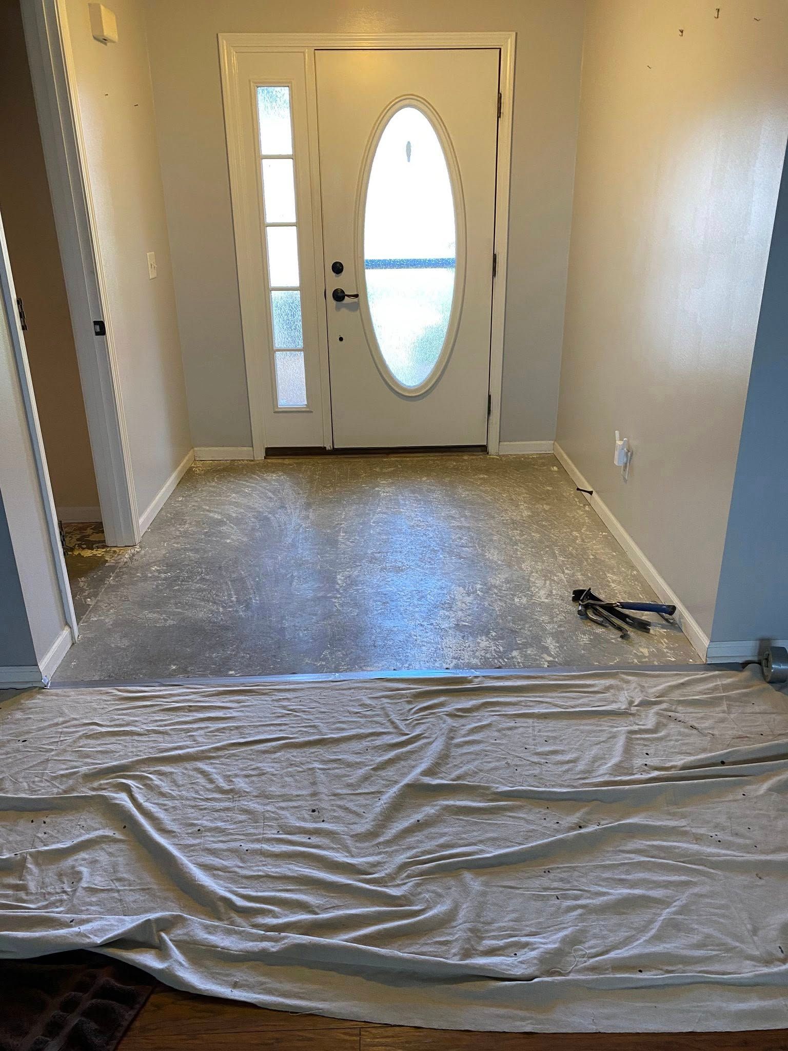 An entryway undergoing renovations, showing a stripped floor and a white front door, with a drop cloth in the foreground.