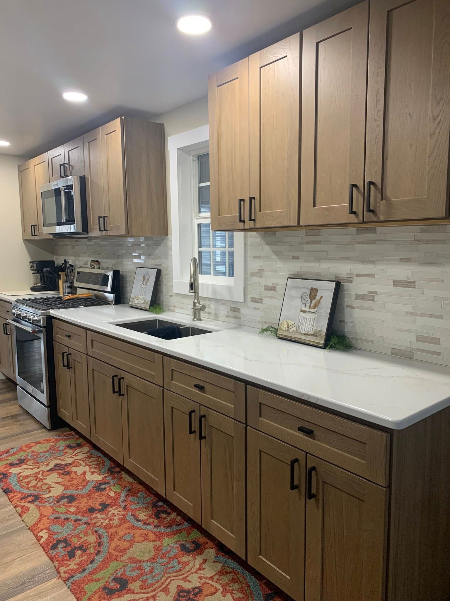 A kitchen with wood-tone cabinets, white countertops, a tiled backsplash, and a patterned rug on a wood floor.