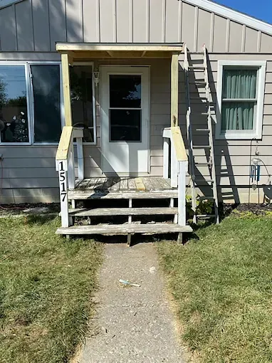 A house entrance with a wood-framed porch, stairs, and a metal ladder leaning against beige siding.