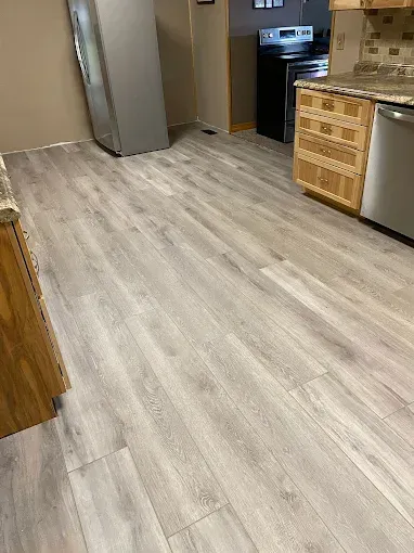 Light gray wood-look laminate flooring installed in a kitchen with wooden cabinets and stainless steel appliances.