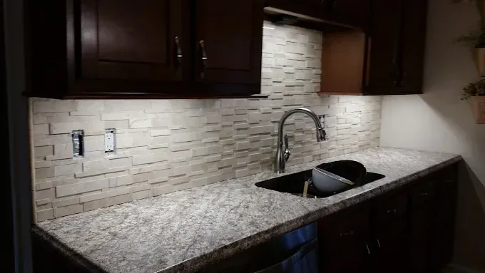 A kitchen sink area with dark brown cabinets, a mottled granite countertop, and a textured, light-colored stone backsplash.