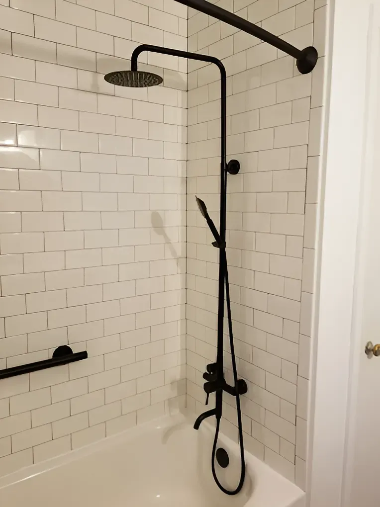 A matte black shower system with a rainfall showerhead, handheld sprayer, and tub faucet mounted on a white subway tile wall.