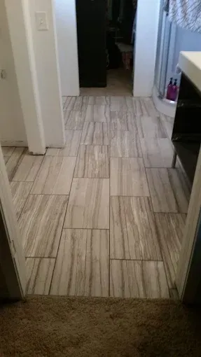 A narrow hallway with light-colored, wood-look rectangular floor tiles leading toward a doorway and bathroom area.