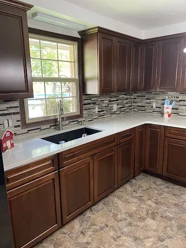 A kitchen with dark wood cabinets, a white countertop, a black farmhouse sink, and a tiled backsplash under a window.