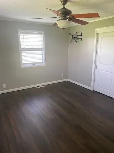 Empty room with gray walls, dark wood-look flooring, a white window, a ceiling fan, and a wall-mounted TV bracket.