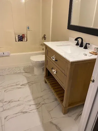 A bathroom featuring a light-wood vanity with a white countertop, marble-patterned flooring, and a tub-shower unit.