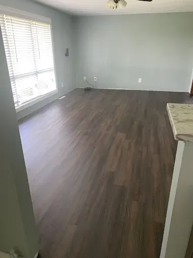 An empty room featuring dark wood-look flooring, light grey walls, and a large window with white horizontal blinds.