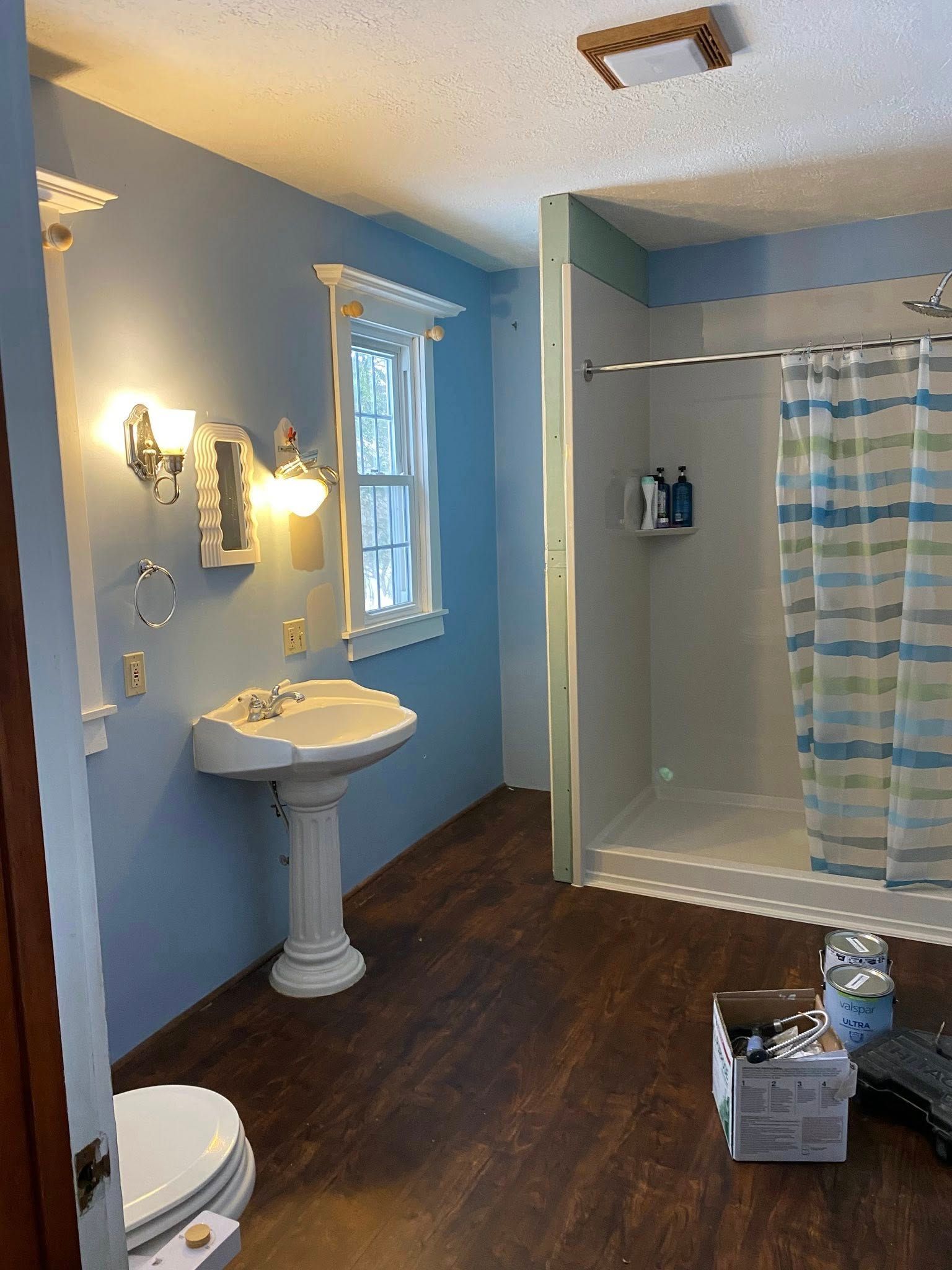 A bathroom with blue walls, a white pedestal sink, a wood-look floor, and a shower with a striped blue and white curtain.