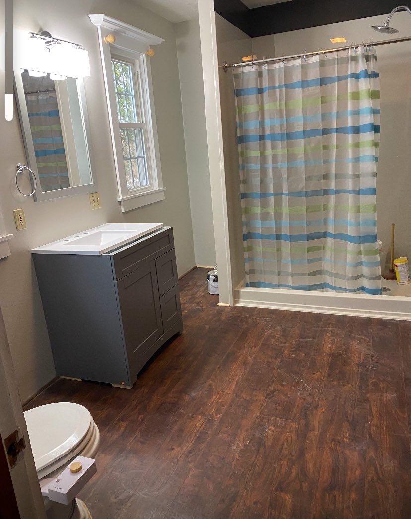 A bathroom featuring a grey vanity with a white sink, a window, a toilet, and a shower with a blue and white striped curtain.