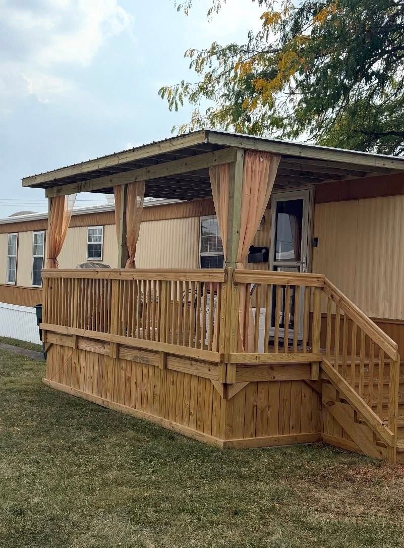 A wooden deck with a metal roof and light-colored curtains attached to a tan mobile home on a grassy lawn.