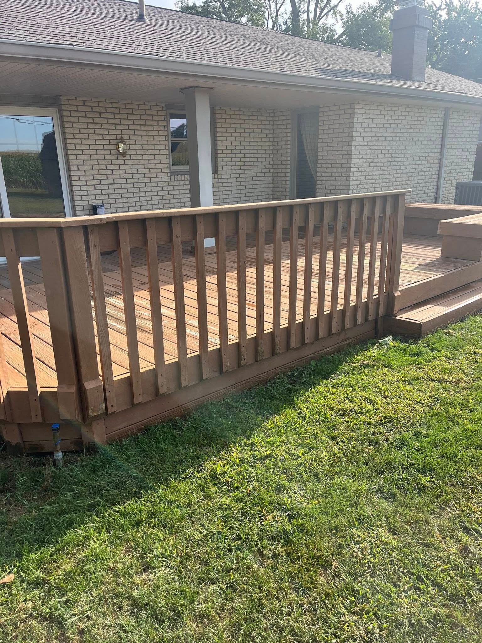 A tan wooden deck with railing attached to the back of a brick house with a gabled roof, viewed from a grassy yard.