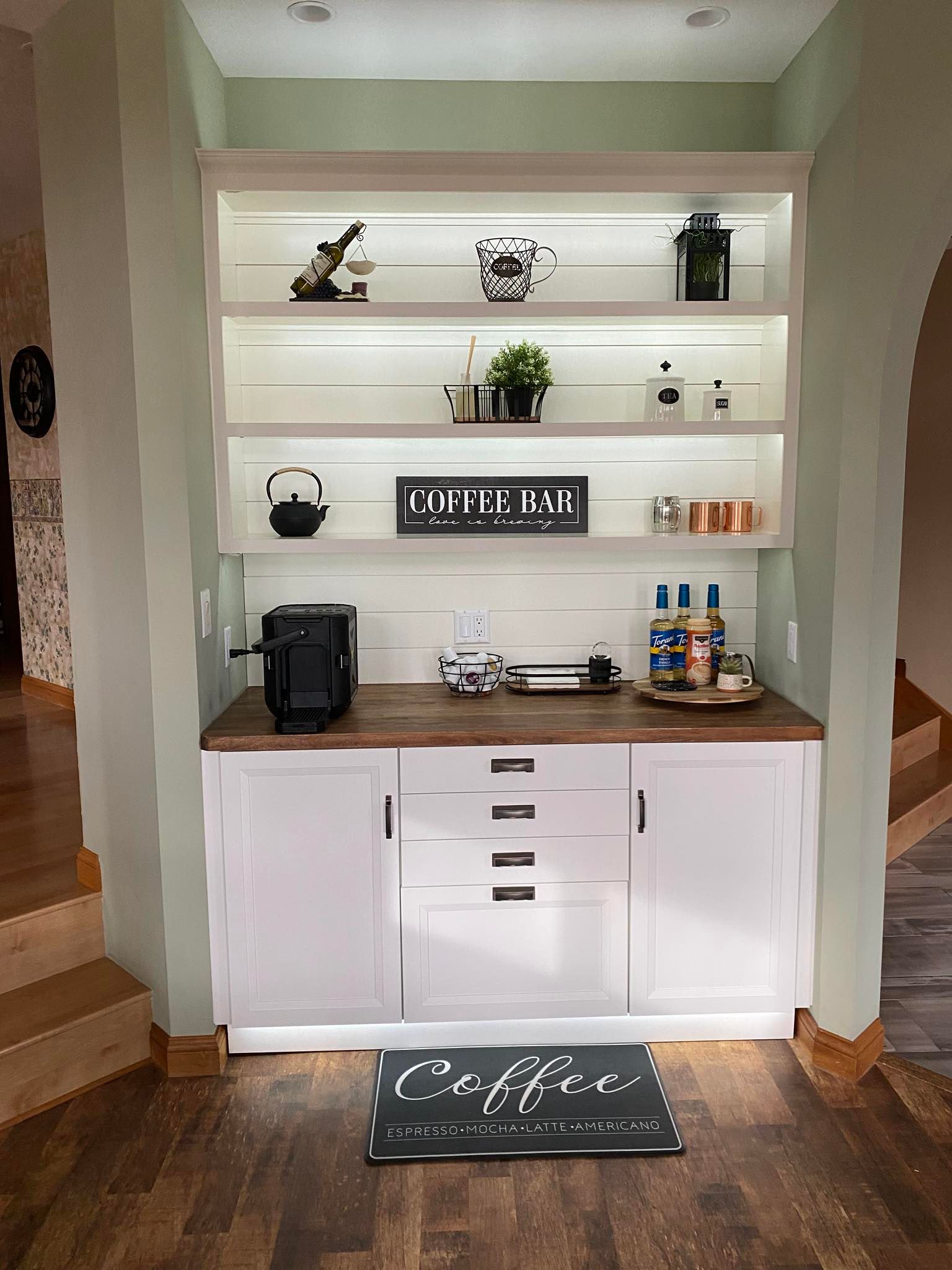 A white built-in coffee station with open shelves, wooden countertop, and base cabinets against light green walls.