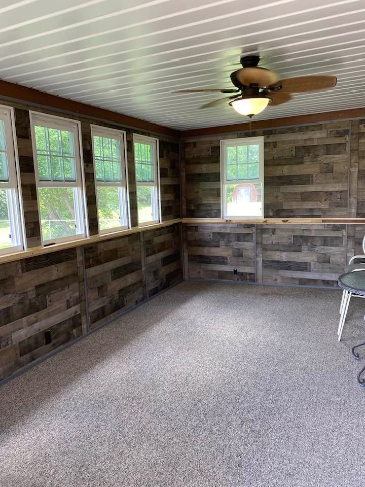 A sunroom with dark reclaimed wood walls, a white ceiling with a ceiling fan, several windows, and speckled flooring.