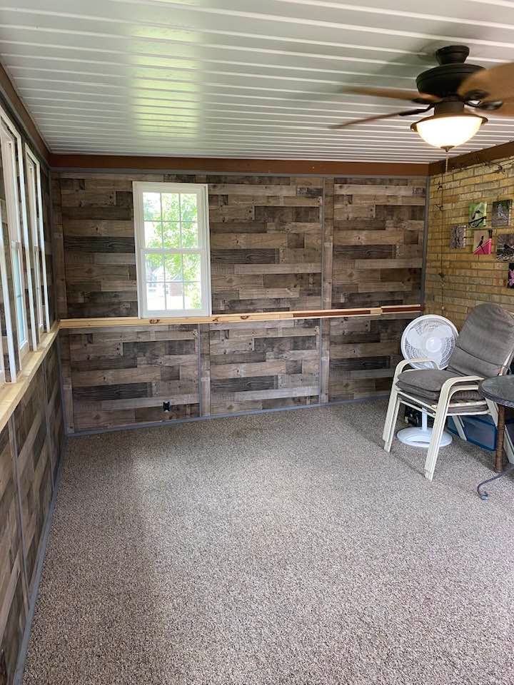 A room with wood-paneled walls, a single window, a ceiling fan, and stacked chairs on a speckled floor.