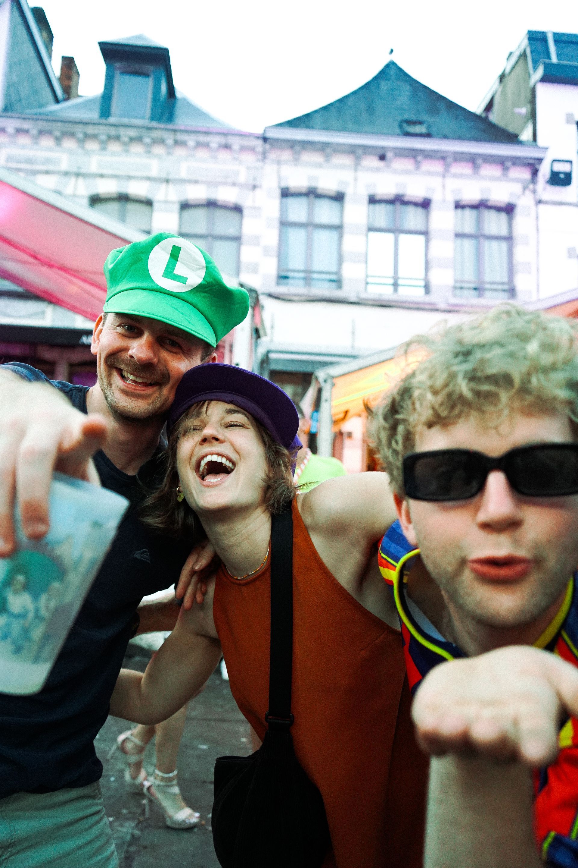 Trois personnes souriantes, l'une portant un chapeau de Luigi, l'autre des lunettes de soleil, s'enlaçant dans la rue.
