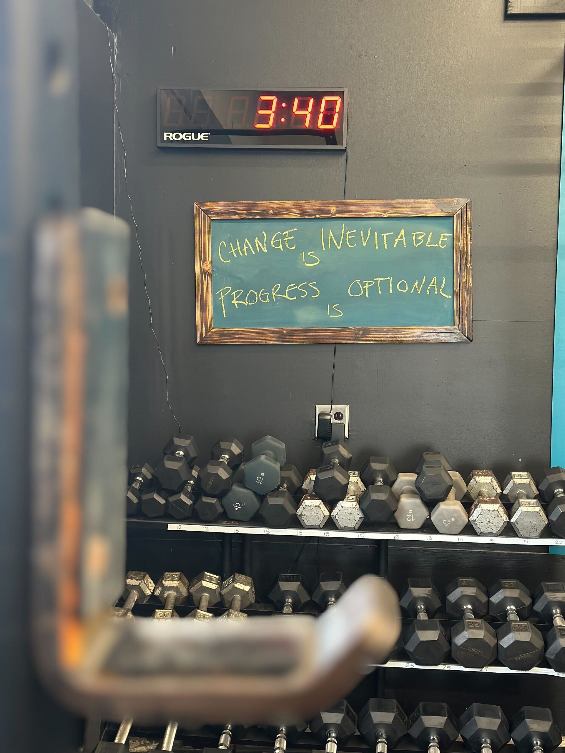 Dumbbell racks in gym below a chalkboard sign and a digital clock displaying 3:40.