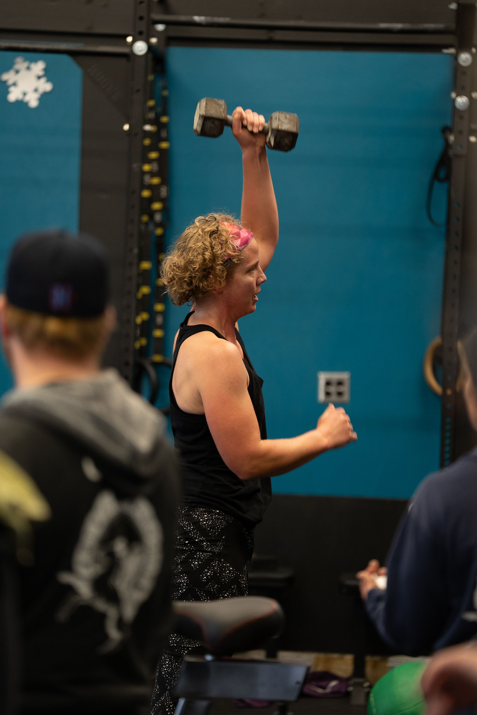 Woman in a gym, lifting a dumbbell overhead. Wearing a black tank top and patterned leggings. Turquoise wall in the background in Portland, OR's best CrossFit gym.