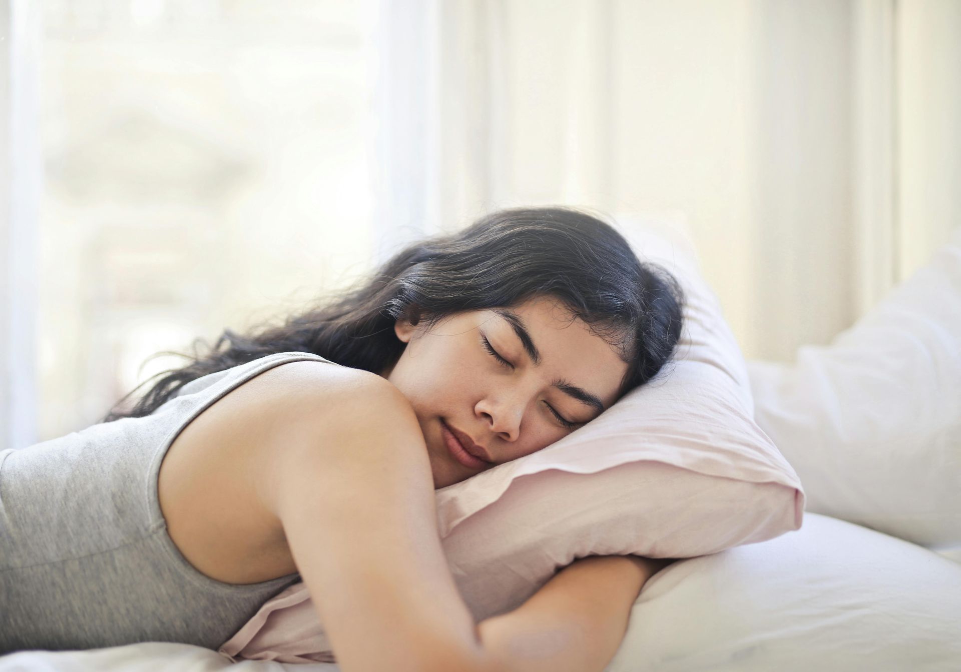 Woman asleep on a bed, head resting on a pillow, near a bright window.