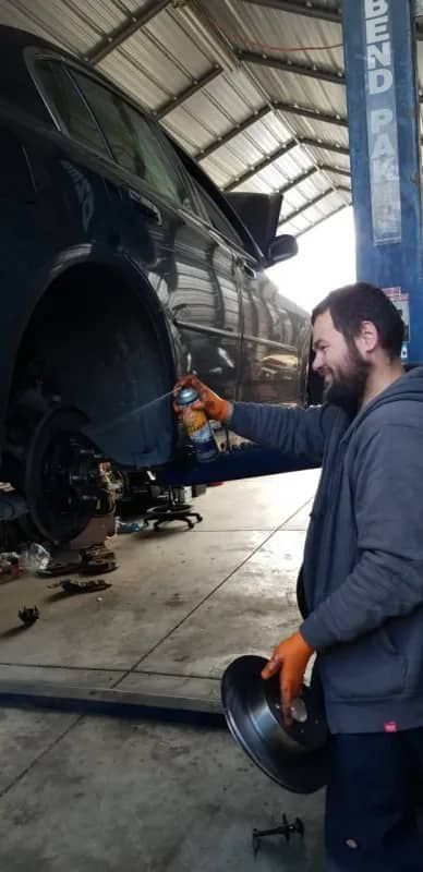 A man in a garage sprays a car part while holding another part. He wears gloves and a grey jacket.
