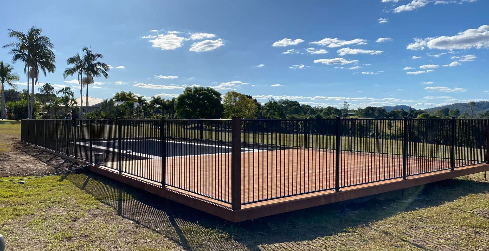 A Swimming Pool with a Wooden Deck and a Glass and Aluminum Fence Surrounding It — RBG Building & Maintenance in Veteran, QLD