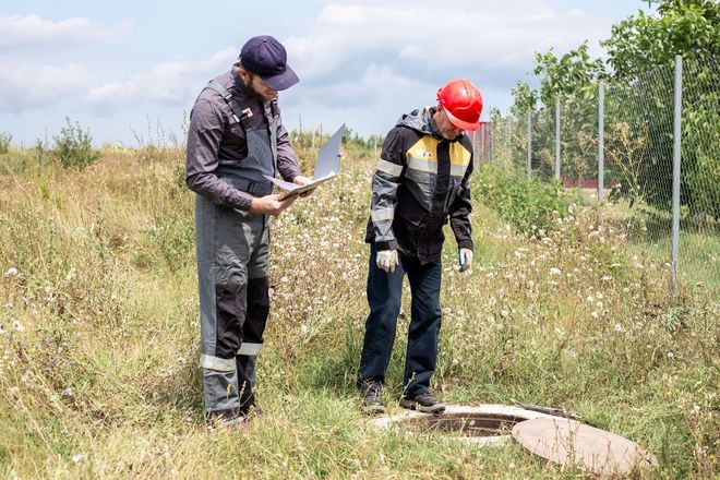 Two workers in safety gear inspect a manhole in a grassy field near a fence.