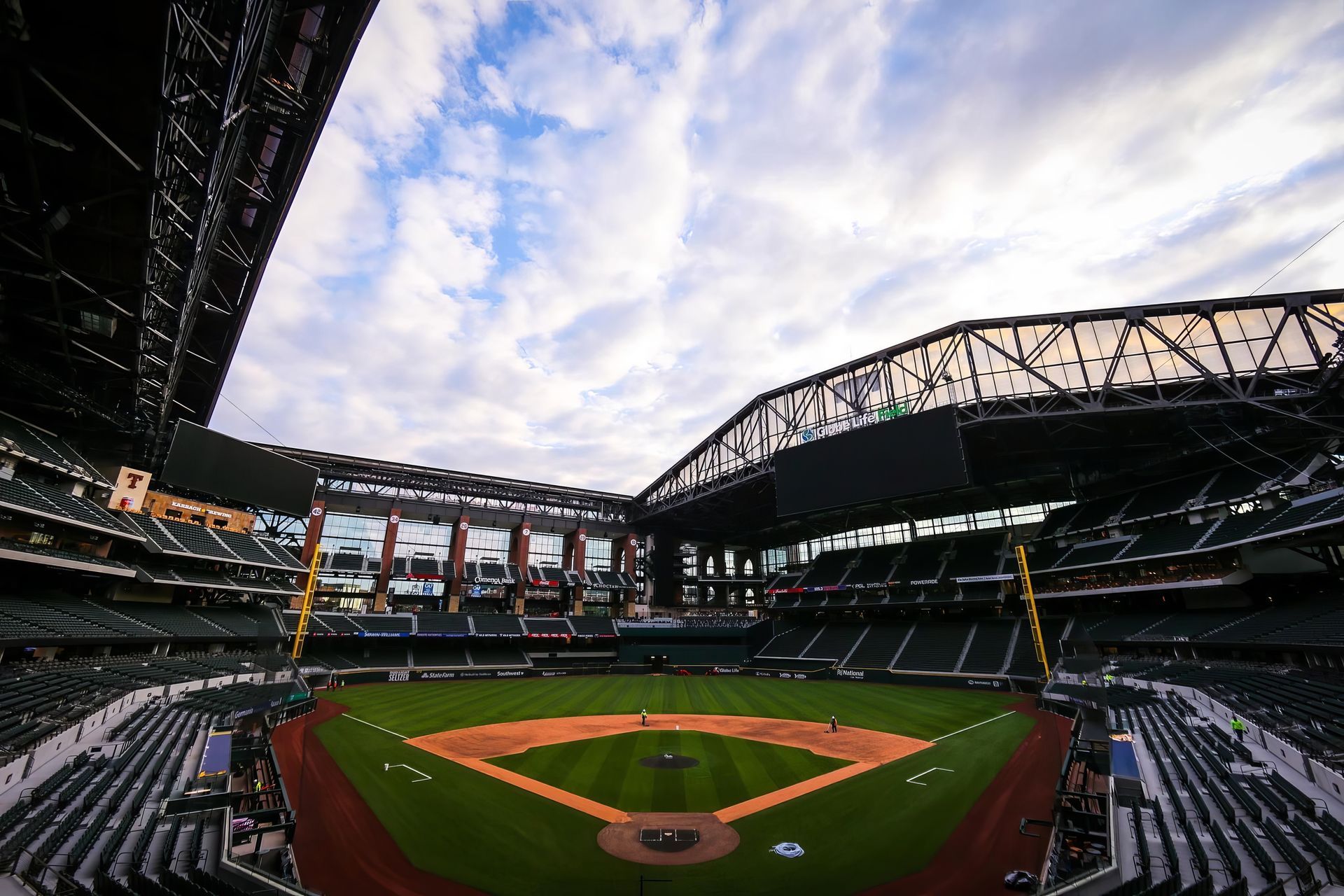 Wide view of an empty baseball stadium with green field and dark seating under a cloudy sky