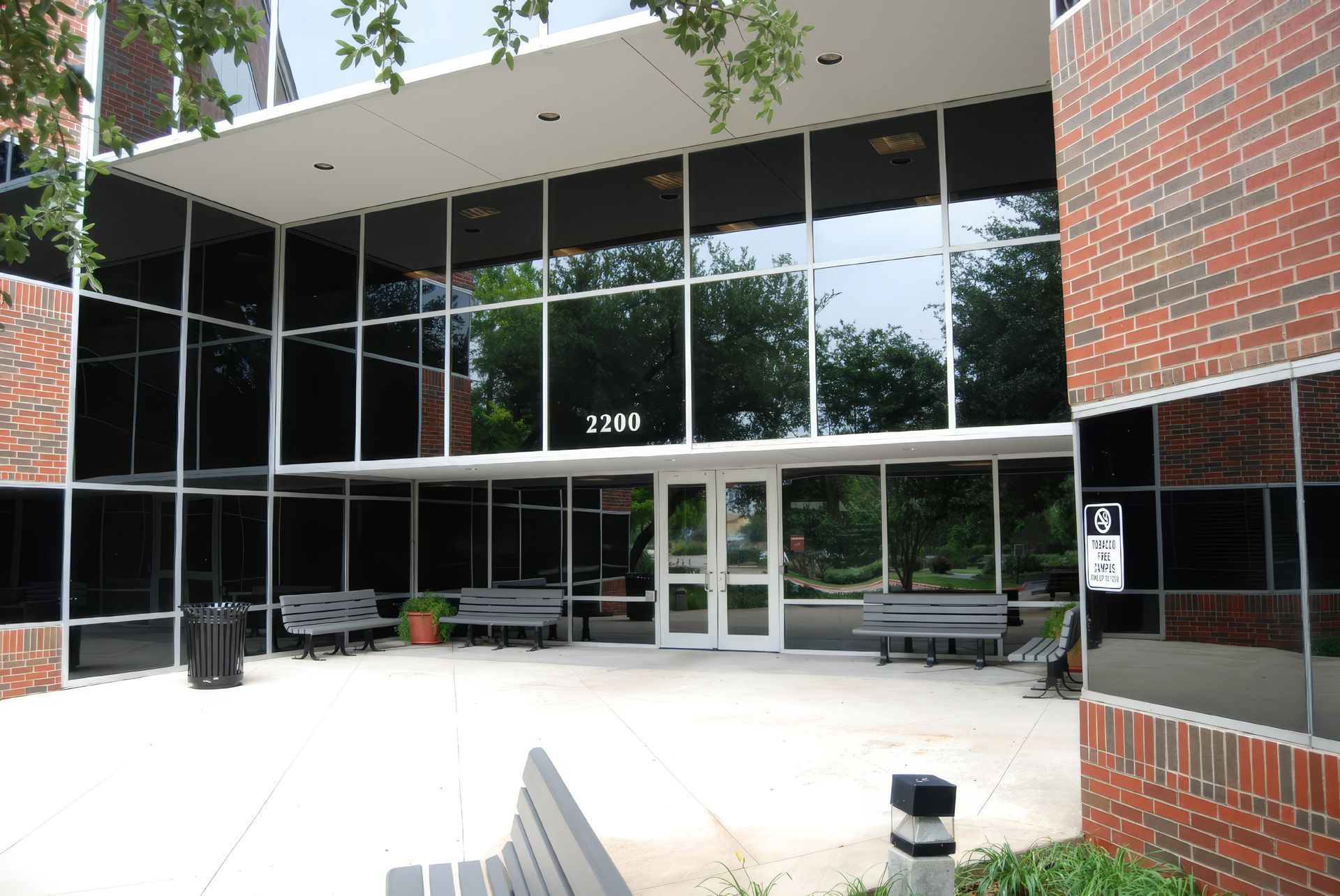 Glass-front building entrance with brick accents, benches, and trees reflected in the windows