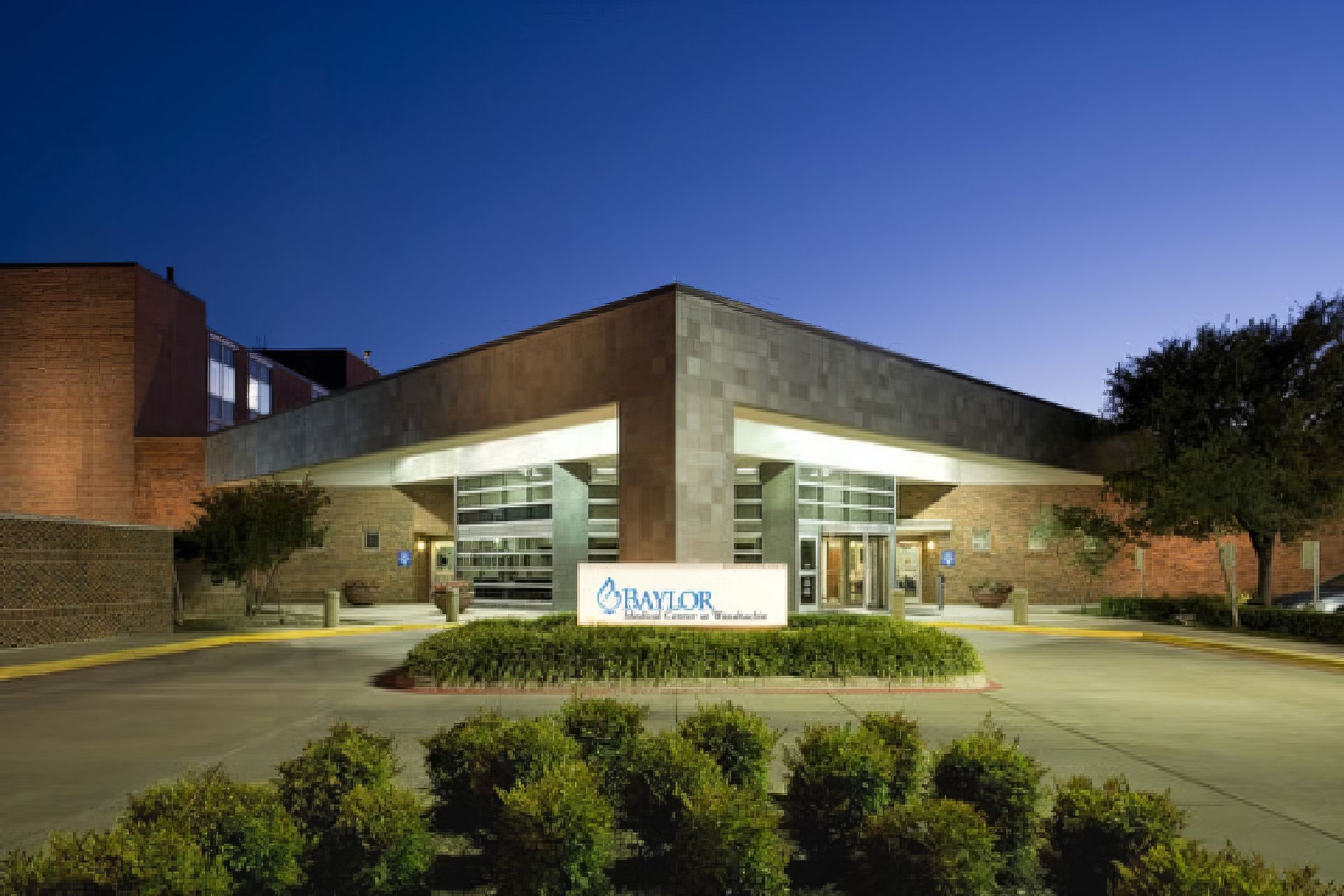 Hospital entrance at dusk with illuminated canopy, driveway, and landscaped bushes.